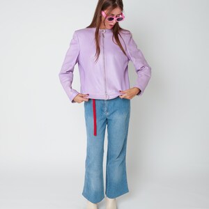 May include: A lavender leather jacket with a full-length silver zipper and a mandarin collar, paired with light blue denim jeans featuring a red fabric belt and cream-colored square-toe boots, accessorized with bright pink oval-frame sunglasses.