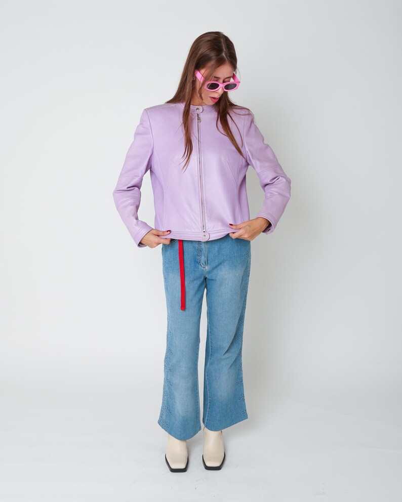 May include: A lavender leather jacket with a full-length silver zipper and a mandarin collar, paired with light blue denim jeans featuring a red fabric belt and cream-colored square-toe boots, accessorized with bright pink oval-frame sunglasses.