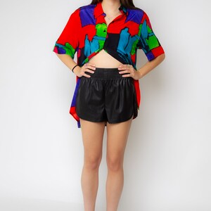 Pode incluir: Uma mulher vestindo uma camisa de manga curta com estampa abstrata colorida, shorts de couro preto e saltos pretos.