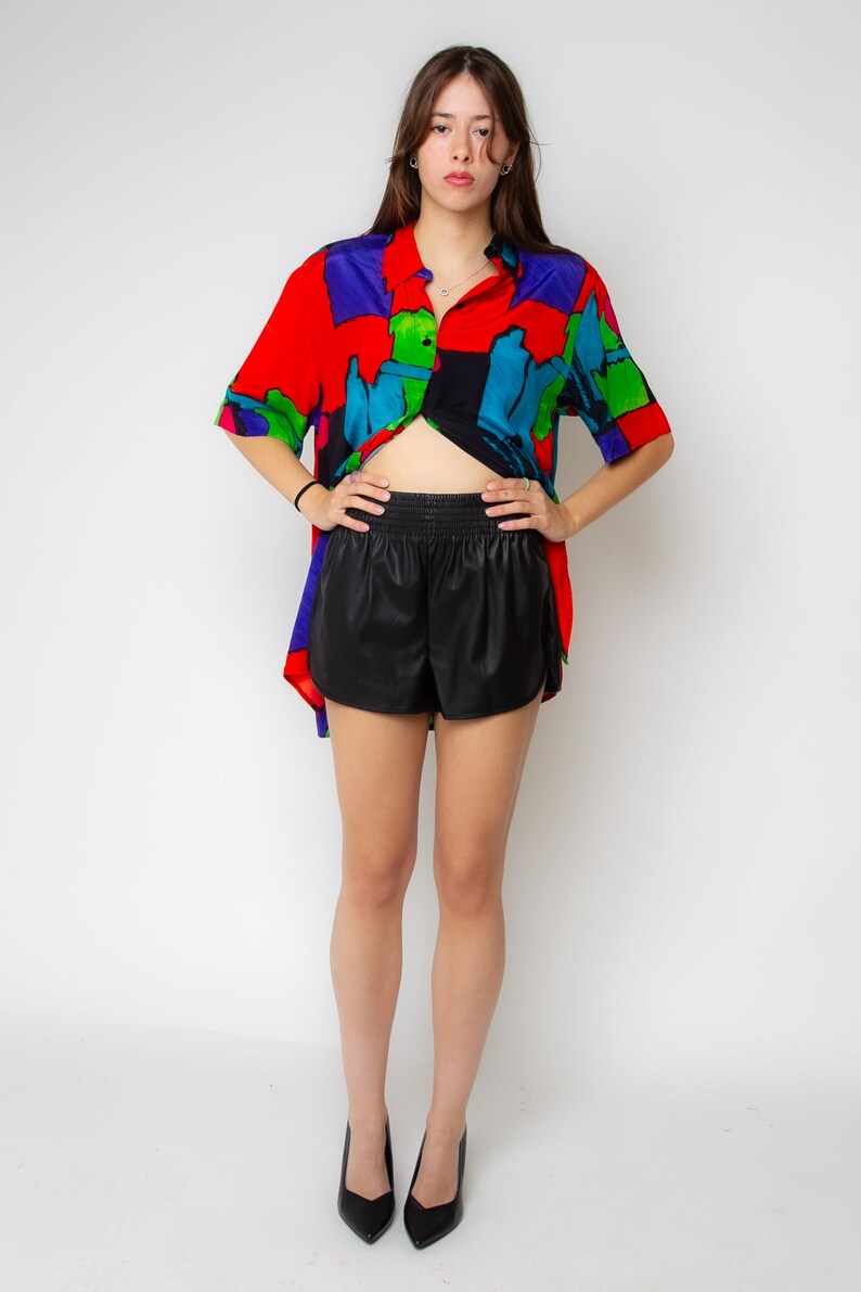 Pode incluir: Uma mulher vestindo uma camisa de manga curta com estampa abstrata colorida, shorts de couro preto e saltos pretos.