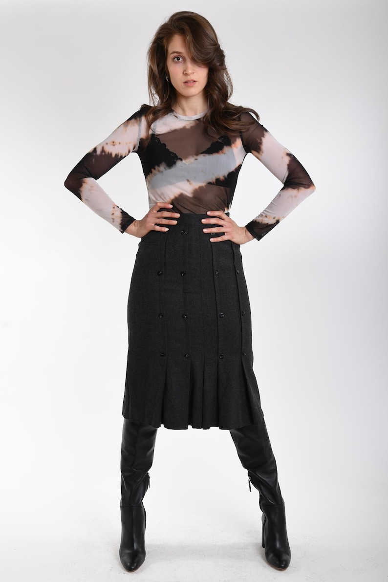 May include: A long-sleeved mesh top with a brown, black, and white tie-dye pattern worn over a black camisole, paired with a dark gray midi skirt featuring vertical pleats and double-breasted buttons, and black knee-high leather boots.