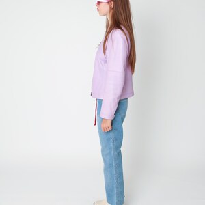 May include: A light purple jacket with long sleeves and a front zipper closure, paired with blue denim jeans featuring a vertical embroidered floral pattern along the side seam and cream-colored ankle boots with a black block heel.