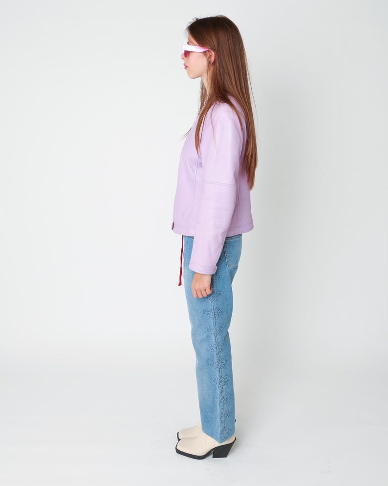 May include: A light purple jacket with long sleeves and a front zipper closure, paired with blue denim jeans featuring a vertical embroidered floral pattern along the side seam and cream-colored ankle boots with a black block heel.