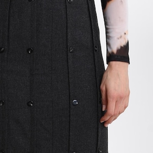 May include: A dark charcoal gray skirt featuring vertical pleats and a series of small, round, matching dark buttons aligned along the pleat lines. The skirt has a high waistband and a structured, tailored appearance with a clean, uniform finish.