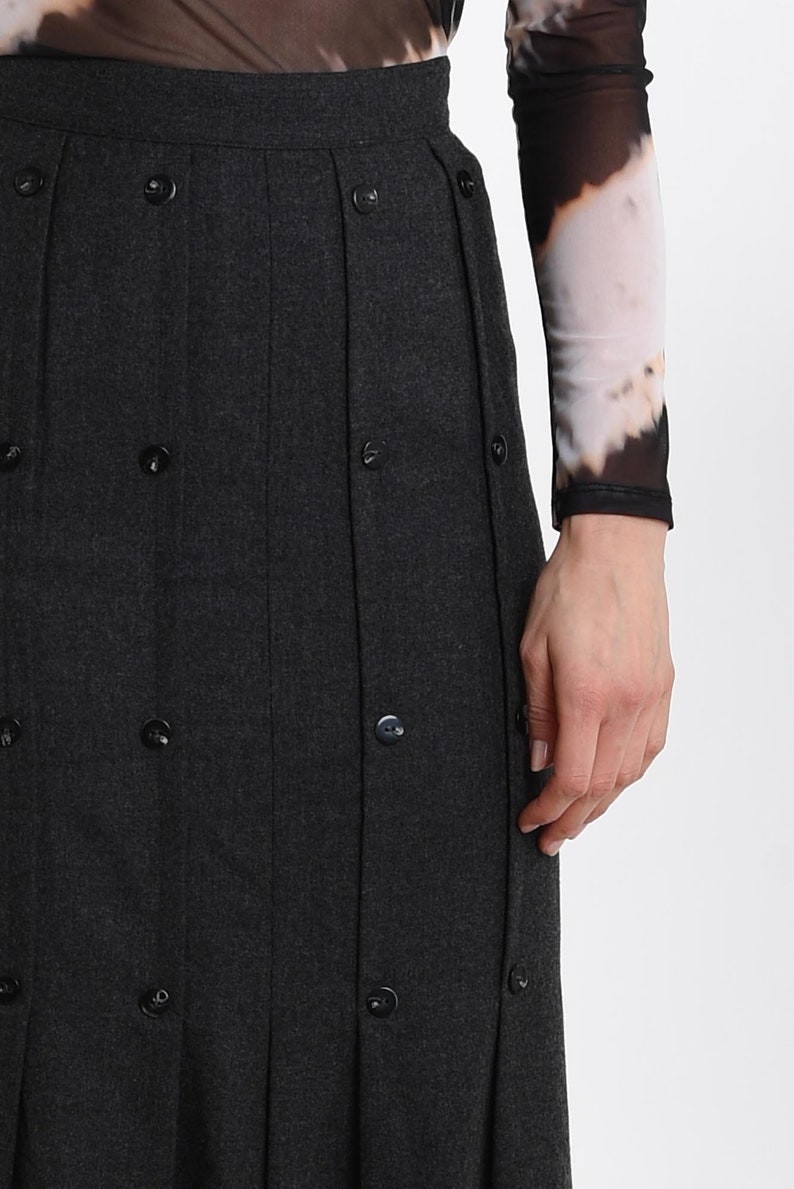 May include: A dark charcoal gray skirt featuring vertical pleats and a series of small, round, matching dark buttons aligned along the pleat lines. The skirt has a high waistband and a structured, tailored appearance with a clean, uniform finish.