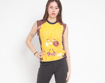 Op de afbeelding: Een mouwloze gele top met een donkerblauwe ronde hals en een grafische print van een gestileerd karaktergezicht met bruin haar, grote ogen en een roze bikinitopje, gecombineerd met een donkergrijze broek.