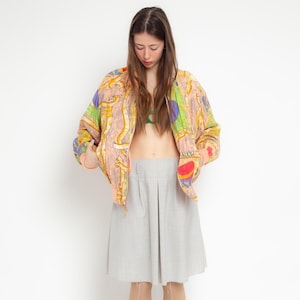 May include: A zip-up bomber jacket featuring a multi-colored abstract print with shades of peach, yellow, green, and purple, paired with a light gray knee-length pleated skirt. The jacket has ribbed cuffs and a visible zipper closure at the front.