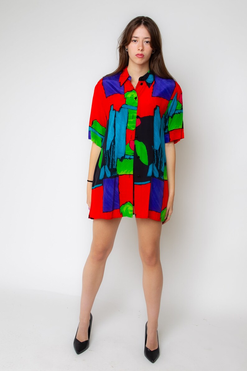 Pode incluir: Uma camisa de bot&otilde;es de manga curta com estampa abstrata em vermelho, azul, verde e preto. A camisa tem um caimento solto e &eacute; feita de um tecido leve.