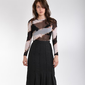 May include: A long-sleeved, sheer mesh top featuring a diagonal tie-dye pattern in black, white, and tan, worn over a black lace bralette. Paired with a charcoal gray, knee-length skirt featuring vertical seams, small black buttons, and pleats.