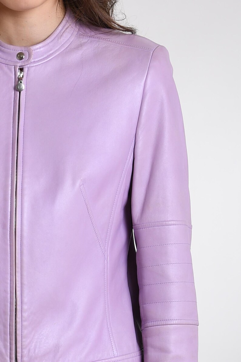 May include: A light purple leather jacket featuring a mandarin collar with a snap button closure, a full-length front zipper, and horizontal stitched detailing along the upper sleeves. The garment has a smooth finish and a fitted silhouette.