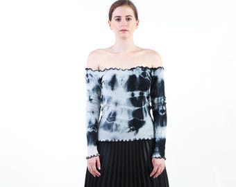 May include: An off-the-shoulder long-sleeve top featuring a black and white tie-dye pattern. The garment has a ribbed texture and a lettuce-edge trim along the neckline, cuffs, and hemline. The top is paired with a black pleated skirt.