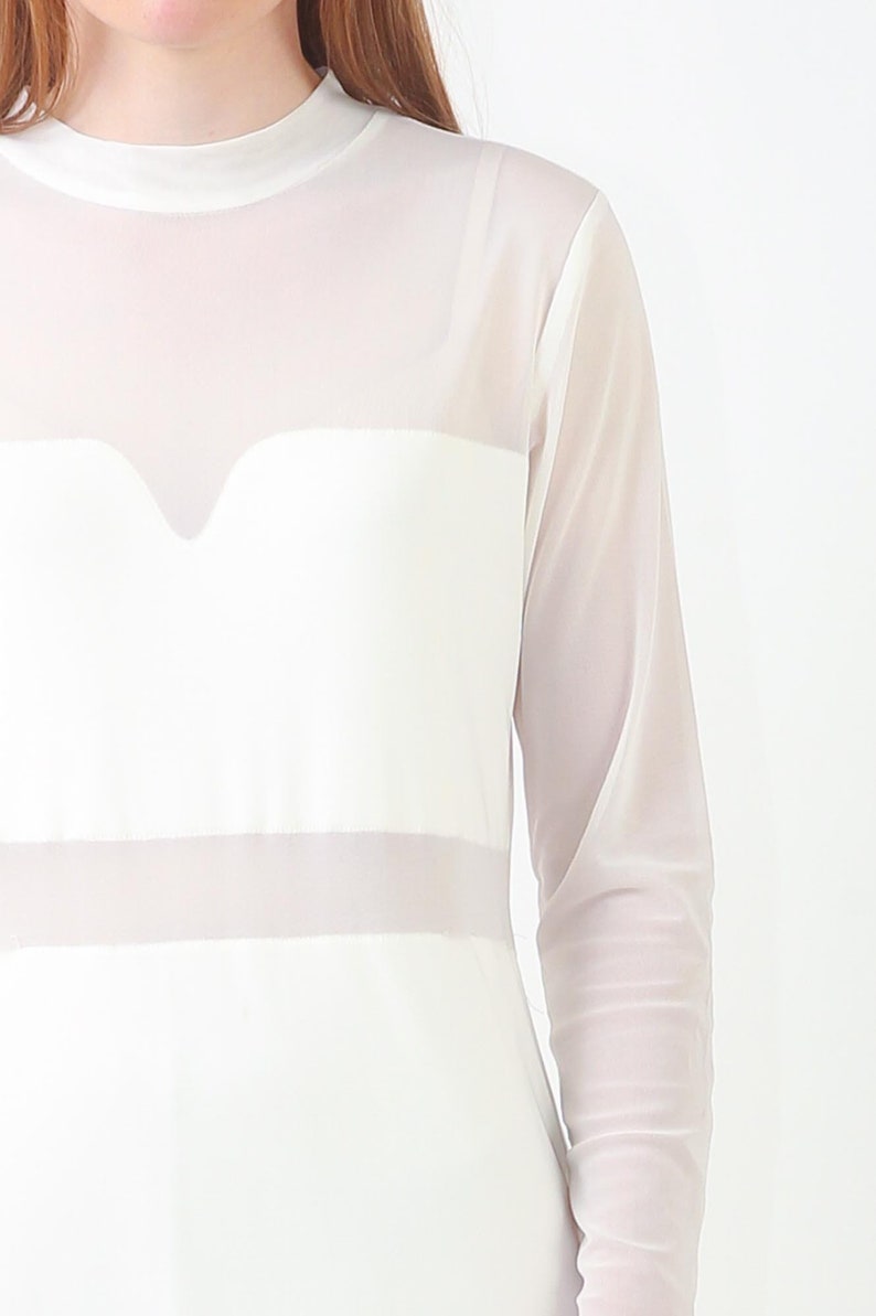 May include: A white long-sleeved top featuring a mock neck and a sheer mesh overlay. The garment includes a solid opaque panel across the chest with a sweetheart-shaped neckline detail and a horizontal sheer band across the midsection.