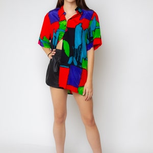 Pode incluir: Uma mulher vestindo uma camisa estampada de vermelho, azul, verde e preto com mangas curtas e uma saia de couro preta. Ela est&aacute; usando sapatos de salto alto pretos.