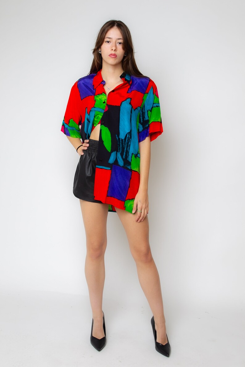 Pode incluir: Uma mulher vestindo uma camisa estampada de vermelho, azul, verde e preto com mangas curtas e uma saia de couro preta. Ela est&aacute; usando sapatos de salto alto pretos.