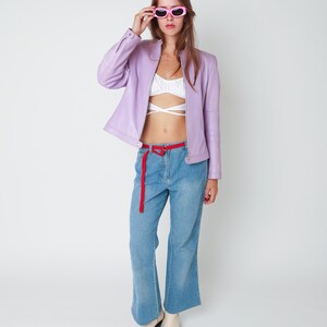 May include: A lavender leather jacket with a front zipper, a white cropped top with crisscross straps, blue denim jeans with a thin red belt, and square-toe cream boots. The outfit is accessorized with bright pink oval-framed sunglasses.