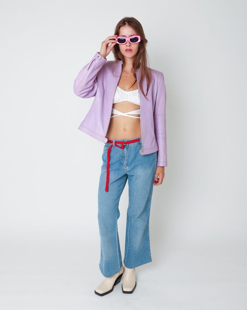 May include: A lavender leather jacket with a front zipper, a white cropped top with crisscross straps, blue denim jeans with a thin red belt, and square-toe cream boots. The outfit is accessorized with bright pink oval-framed sunglasses.