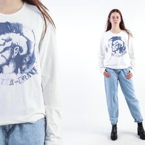 Könnte beinhalten: Weißes Langarm-T-Shirt mit einem blauen Grafikdruck eines stilisierten Indianerkopfes mit Irokesenschnitt und dem Text "Only The Brave" in einem Kreis um den Kopf.