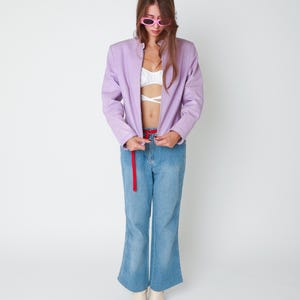 May include: A lavender zip-up jacket worn over a white cropped top, paired with light blue denim jeans featuring a red belt and cream-colored square-toe boots. The outfit is accessorized with pink oval-framed sunglasses.