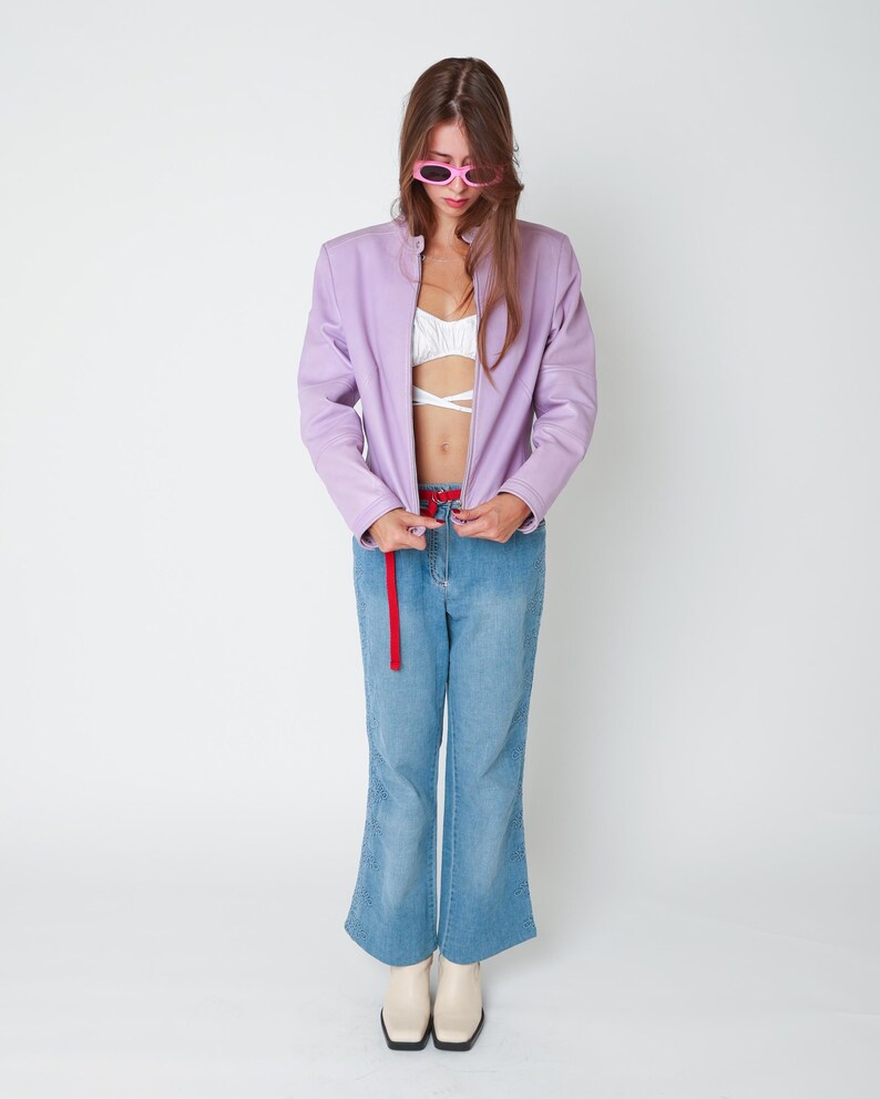 May include: A lavender zip-up jacket worn over a white cropped top, paired with light blue denim jeans featuring a red belt and cream-colored square-toe boots. The outfit is accessorized with pink oval-framed sunglasses.