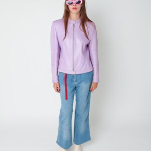 May include: A lavender leather jacket with a mandarin collar and a full-length front zipper, paired with light blue denim jeans featuring a red fabric belt and side-leg cutouts, finished with off-white ankle boots and pink-framed sunglasses.