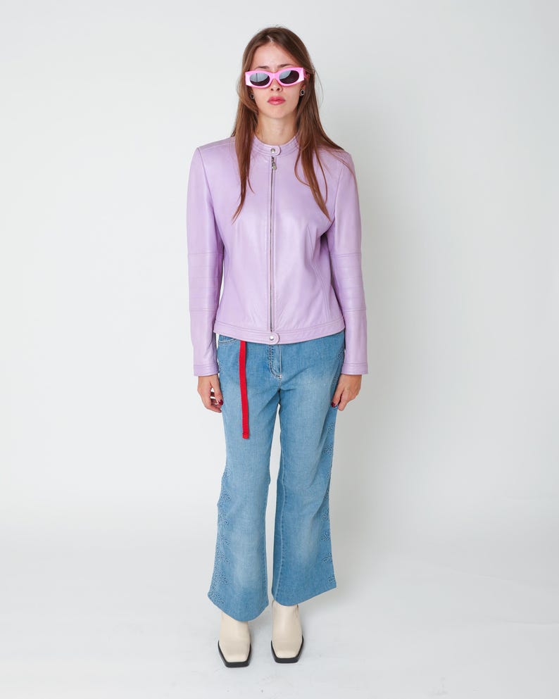 May include: A lavender leather jacket with a mandarin collar and a full-length front zipper, paired with light blue denim jeans featuring a red fabric belt and side-leg cutouts, finished with off-white ankle boots and pink-framed sunglasses.