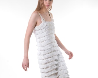 May include: A white sleeveless dress featuring multiple horizontal tiers of fringe covering the entire length of the garment. The dress has two shoulder straps, each accented with a circular floral-like embellishment at the top of the bodice.