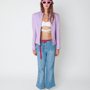 May include: A lavender leather jacket, a white cropped wrap top, light blue denim jeans with side embroidery, a thin red belt, and pink sunglasses. The outfit is completed with cream-colored square-toe boots, creating a layered look.