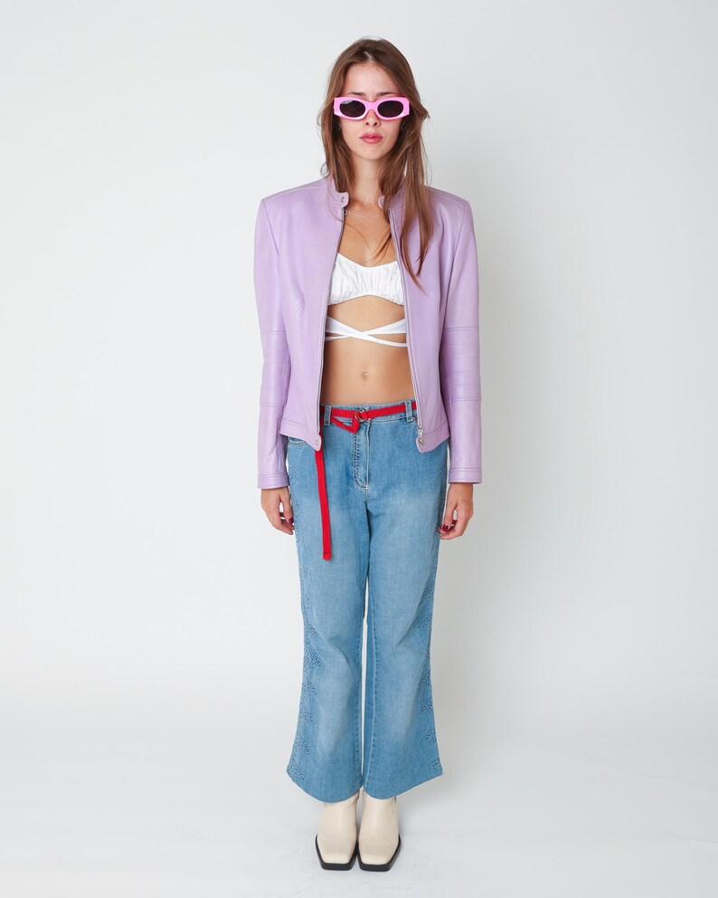 May include: A lavender leather jacket, a white cropped wrap top, light blue denim jeans with side embroidery, a thin red belt, and pink sunglasses. The outfit is completed with cream-colored square-toe boots, creating a layered look.