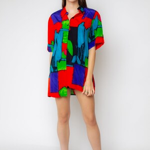 Pode incluir: Uma mulher vestindo uma camisa de bot&atilde;o de manga curta com uma estampa abstrata colorida. A camisa &eacute; vermelha, azul, verde e preta. Ela est&aacute; usando sapatos de salto alto pretos.