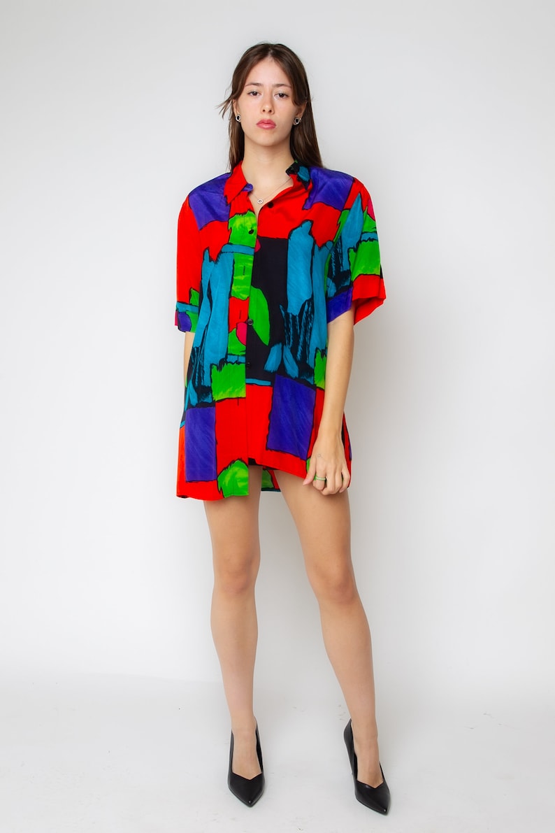 Pode incluir: Uma mulher vestindo uma camisa de bot&atilde;o de manga curta com uma estampa abstrata colorida. A camisa &eacute; vermelha, azul, verde e preta. Ela est&aacute; usando sapatos de salto alto pretos.