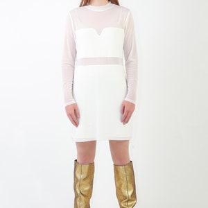 May include: A white long-sleeved dress featuring a sheer mesh overlay on the upper chest and sleeves, paired with knee-high metallic gold boots. The dress has a high neckline and a solid opaque white bodice section with a sweetheart-style neckline.