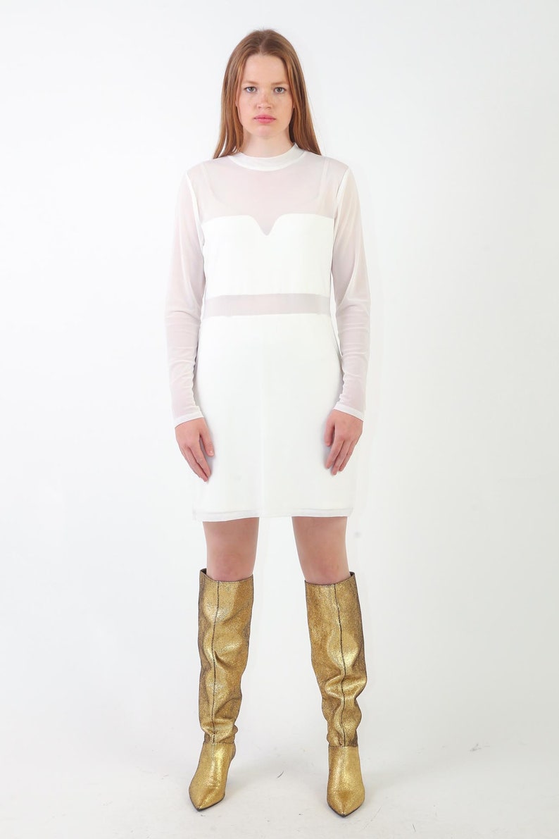 May include: A white long-sleeved dress featuring a sheer mesh overlay on the upper chest and sleeves, paired with knee-high metallic gold boots. The dress has a high neckline and a solid opaque white bodice section with a sweetheart-style neckline.