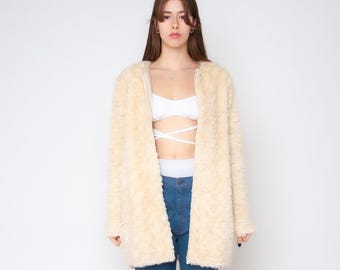 Puede incluir: Una chaqueta de piel sintética color crema, de manga larga y frente abierto, con un acabado texturizado de pelo largo. La prenda se lleva sobre un top corto blanco tipo bralette con tiras y unos vaqueros azules de tiro alto con costuras.