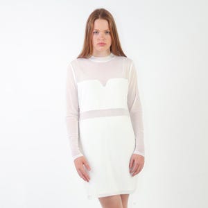 May include: A white long-sleeve dress featuring a sheer mesh overlay on the upper chest and sleeves, a high neckline, and a solid opaque white panel across the bodice and skirt. The garment has a straight silhouette and a clean, minimalist design.