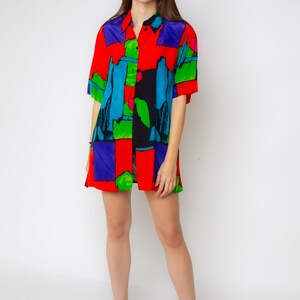 Pode incluir: Uma camisa de bot&otilde;es de manga curta com uma estampa abstrata colorida em tons de vermelho, azul, verde e preto. A camisa tem um caimento solto e uma gola.