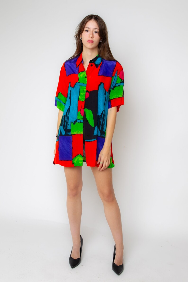 Pode incluir: Uma camisa de bot&otilde;es de manga curta com uma estampa abstrata colorida em tons de vermelho, azul, verde e preto. A camisa tem um caimento solto e uma gola.
