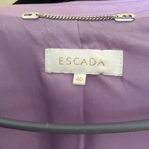 May include: A lavender jacket interior featuring a white fabric label with the text ESCADA in gold lettering and a smaller tag below with the number 40. A silver-toned metal chain loop is attached at the neckline above the brand label.