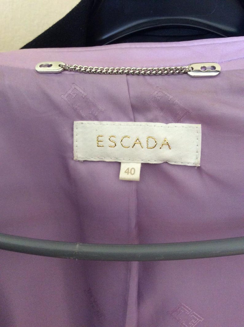 May include: A lavender jacket interior featuring a white fabric label with the text ESCADA in gold lettering and a smaller tag below with the number 40. A silver-toned metal chain loop is attached at the neckline above the brand label.