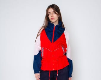 May include: A color-blocked windbreaker jacket featuring a vibrant red central panel, a dark blue high collar and lower sleeve sections, and light pink upper sleeves. The jacket includes a front zipper closure and a drawstring waist with red cords.
