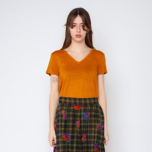 Könnte beinhalten: Ein senfgelbes Kurzarm-T-Shirt mit V-Ausschnitt kombiniert mit einem dunkelgrün-schwarz karierten Rock, der mit kleinen, verstreuten, mehrfarbigen Blumenstickereien über den gesamten Stoff versehen ist und einen deutlichen Kontrast bildet.
