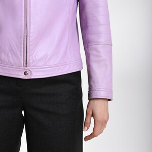 May include: A light purple leather jacket featuring a central silver-toned zipper, a snap button closure at the hem, and horizontal stitched detailing across the sleeves, paired with dark charcoal gray trousers.