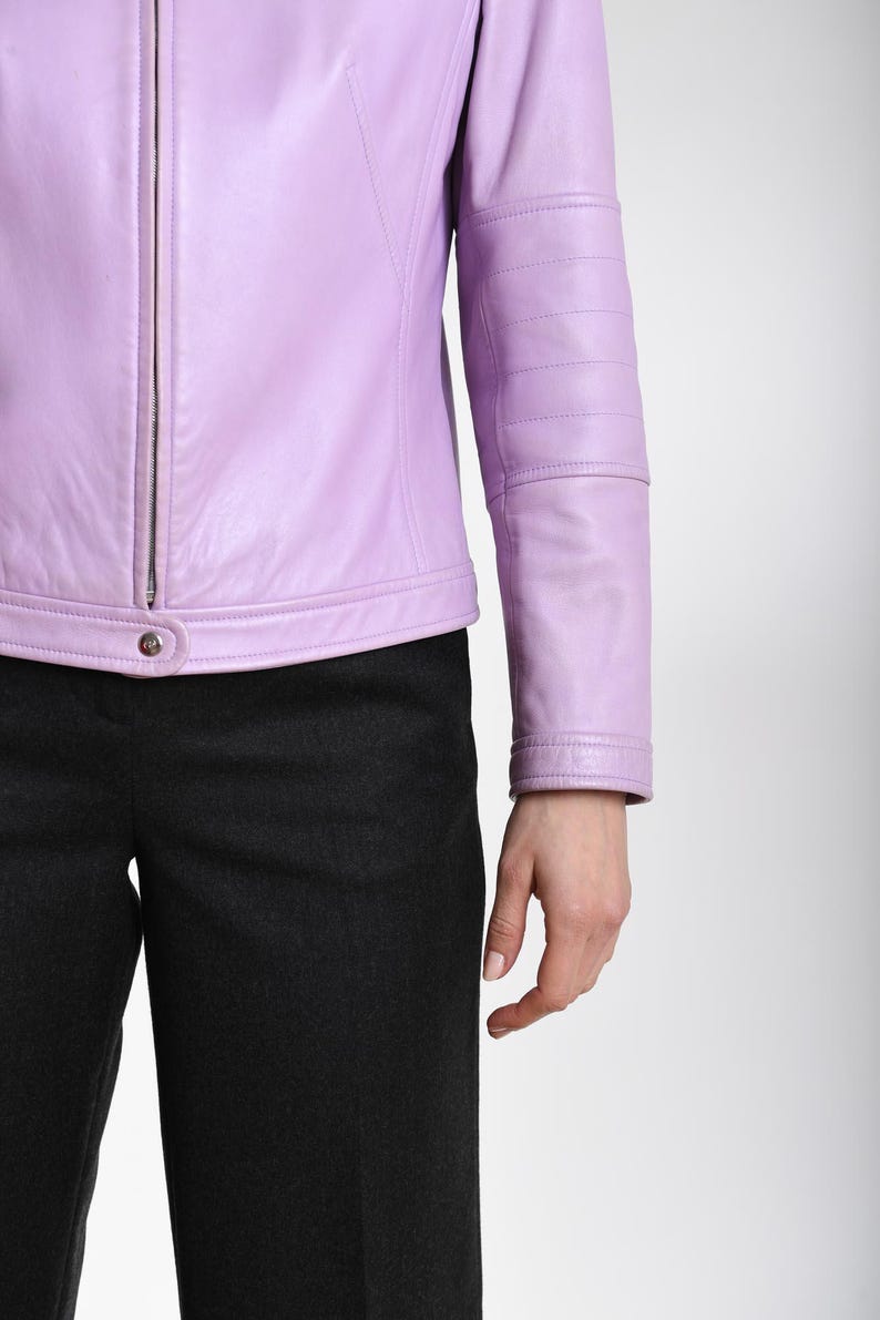 May include: A light purple leather jacket featuring a central silver-toned zipper, a snap button closure at the hem, and horizontal stitched detailing across the sleeves, paired with dark charcoal gray trousers.