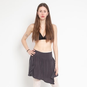 May include: A dark gray mini skirt featuring an asymmetrical hemline with ruffled detailing along the sides and a smooth waistband. The skirt is paired with a black triangle top and white patterned leggings, creating a layered outfit.