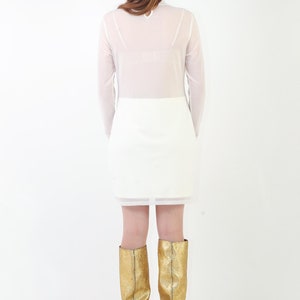 May include: A long-sleeved white dress featuring a sheer upper bodice and a solid opaque skirt section. The dress is paired with knee-high metallic gold boots that have a textured finish and a small heel, viewed from the back against a white background.