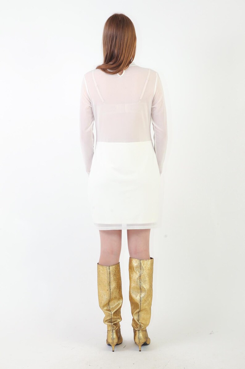 May include: A long-sleeved white dress featuring a sheer upper bodice and a solid opaque skirt section. The dress is paired with knee-high metallic gold boots that have a textured finish and a small heel, viewed from the back against a white background.