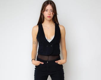 May include: A black sleeveless vest featuring a deep V-neckline and a double-breasted front with two visible buttons. The garment is cropped at the waist and is worn over a white lace-trimmed camisole and black trousers with a sheer black midsection.