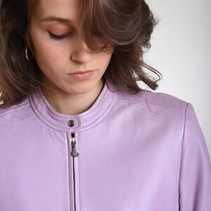 May include: A light purple leather jacket featuring a mandarin collar with a single snap button closure and a central silver-toned metal zipper. The jacket has a smooth, matte finish and long sleeves, providing a clean and structured appearance.