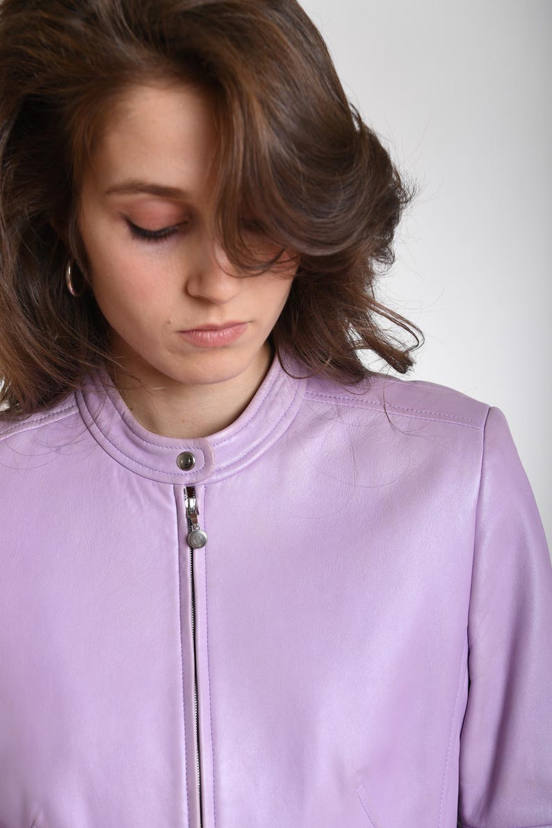 May include: A light purple leather jacket featuring a mandarin collar with a single snap button closure and a central silver-toned metal zipper. The jacket has a smooth, matte finish and long sleeves, providing a clean and structured appearance.