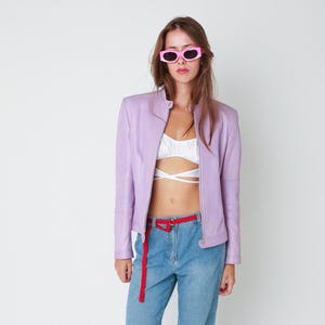 May include: A light purple leather jacket with a stand-up collar and a front zipper, worn over a white cropped top with crisscross straps, paired with blue denim jeans featuring a red fabric belt and bright pink rectangular sunglasses.