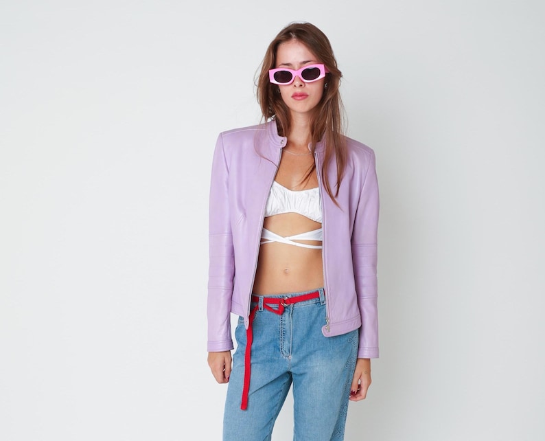 May include: A light purple leather jacket with a stand-up collar and a front zipper, worn over a white cropped top with crisscross straps, paired with blue denim jeans featuring a red fabric belt and bright pink rectangular sunglasses.
